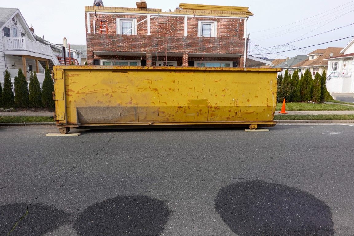 Large yellow dumpster on a residential street in front of a brick house, with dark wet patches on the pavement.