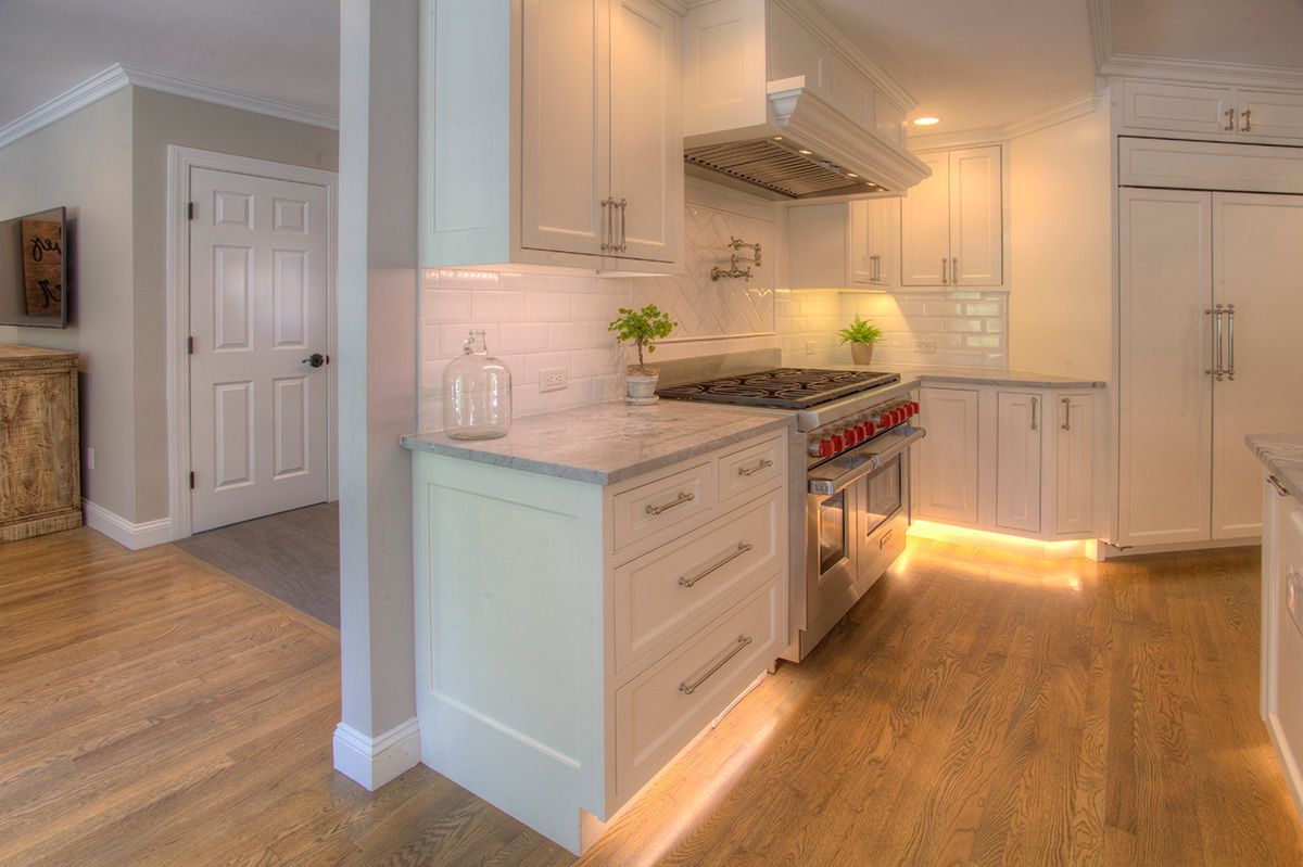 A well-lit white kitchen with light wood floors, featuring cabinets, a stove, and under-cabinet lighting.