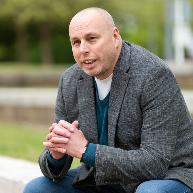 Man in gray blazer seated outdoors, leaning forward with hands clasped and looking at the camera.