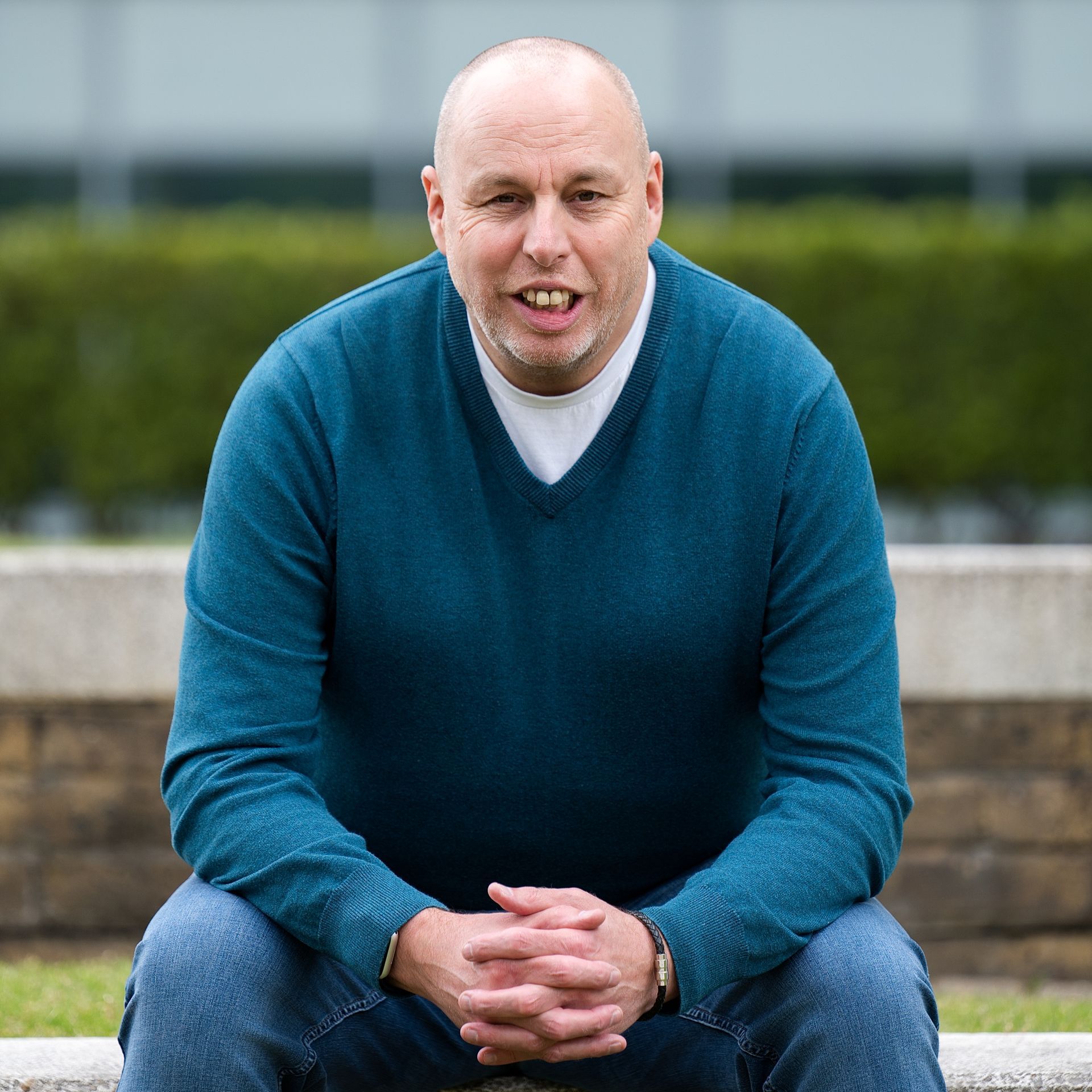 Man sitting on a stone bench outdoors, wearing a teal sweater and jeans, with a hedge behind him.