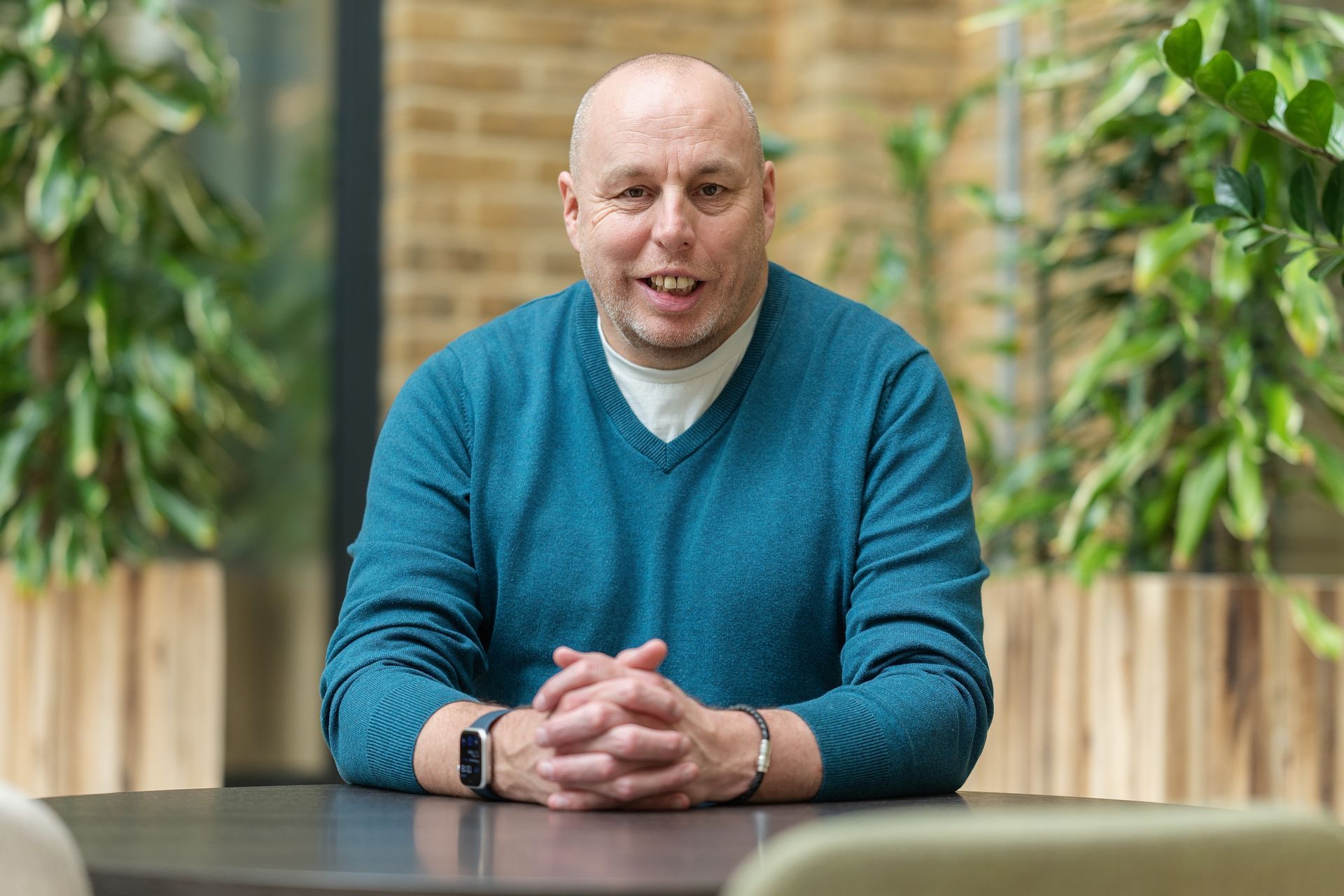 Man in a teal sweater sits at a table in a bright indoor space with plants and brick walls.