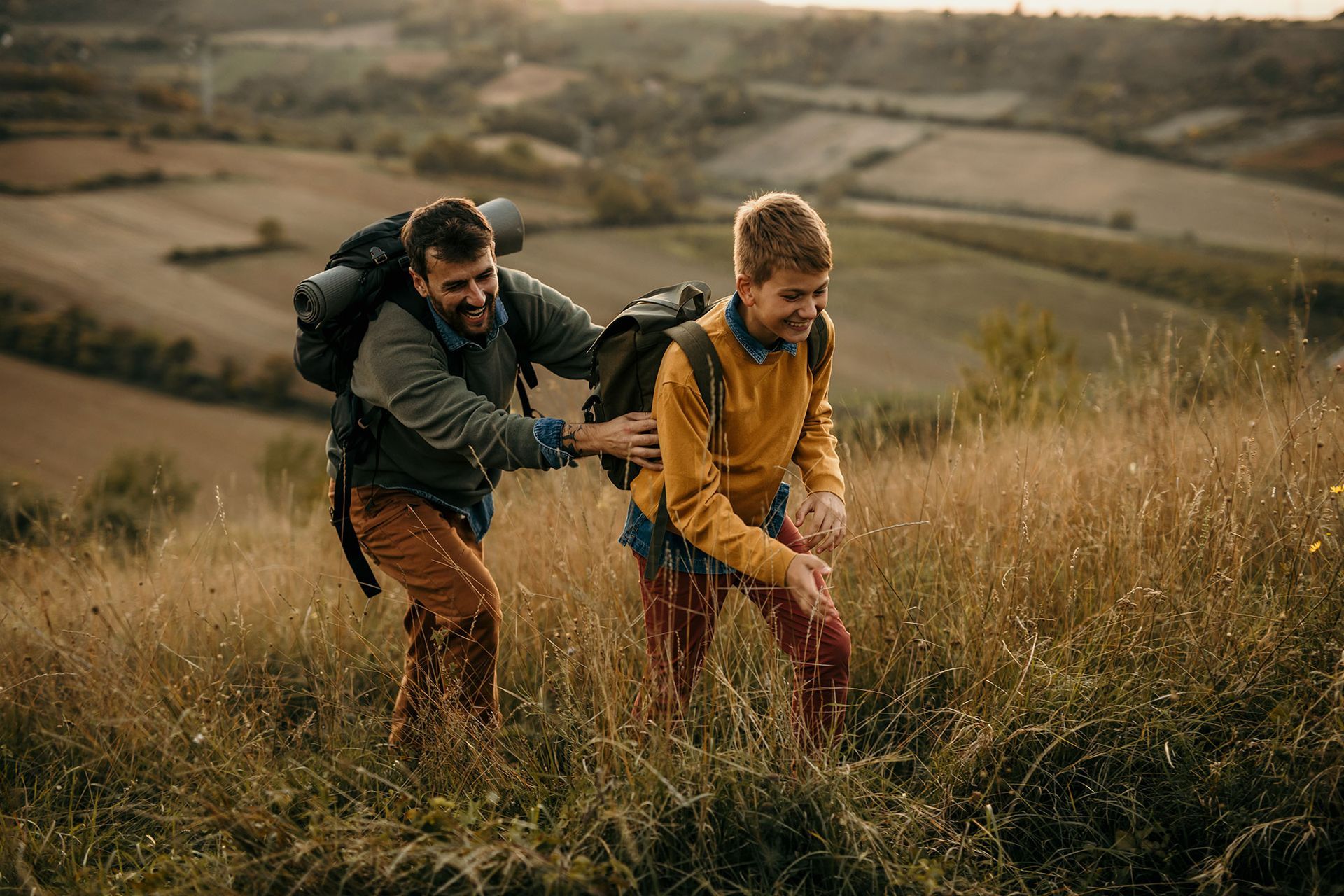 Two hikers with backpacks walking through tall grass in a rural field at sunset