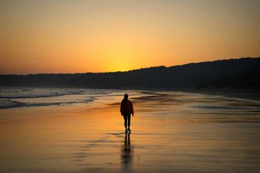 Person walking on a reflective beach at sunset with orange sky and dark shoreline