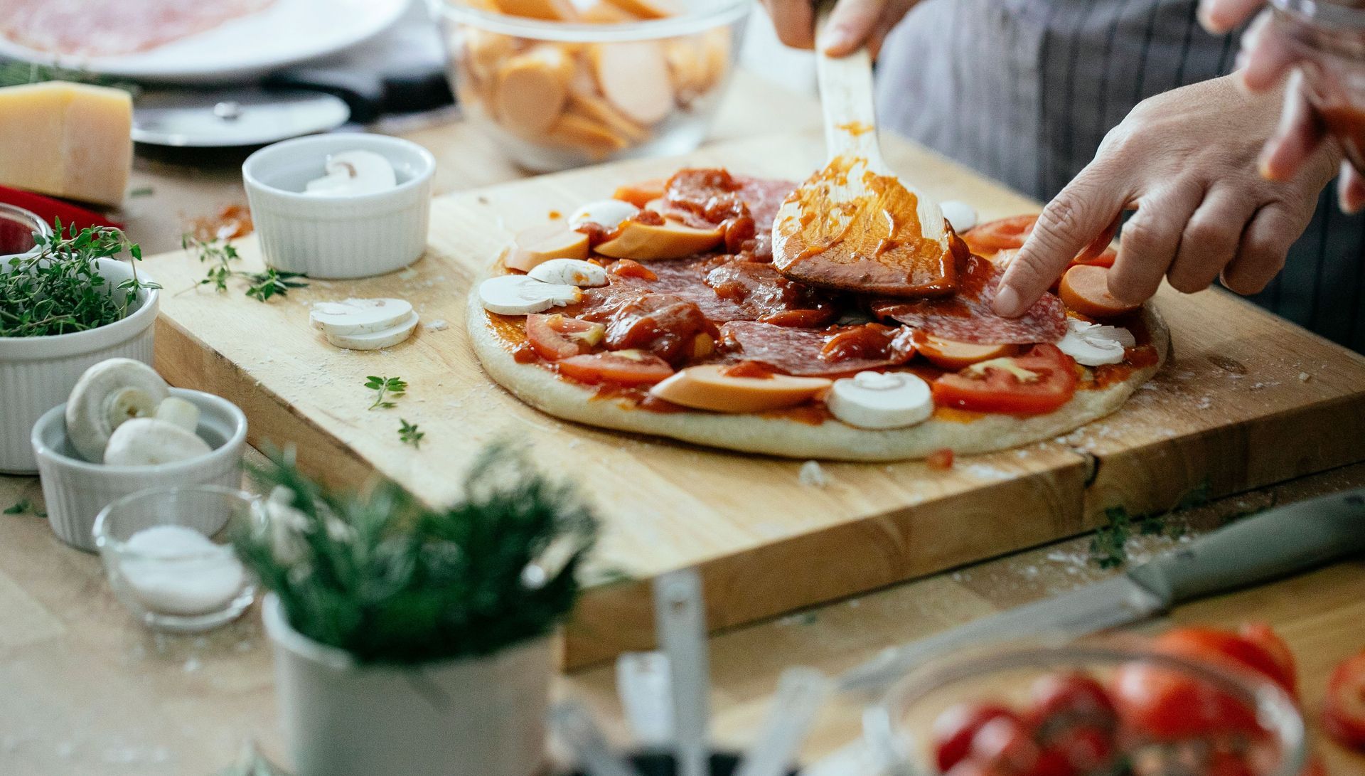 Hands cutting a pepperoni pizza on a wooden board with sauces and herbs nearby.