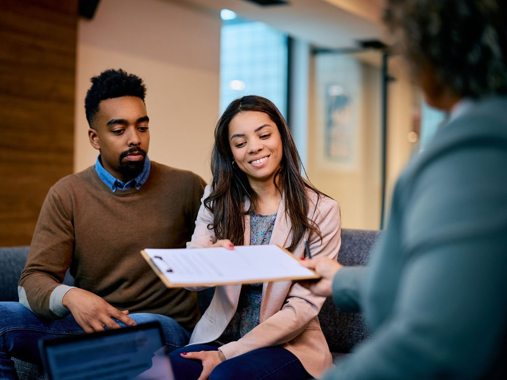 Smiling woman takes a clipboard with documents while sitting next to a man in a modern office setting.