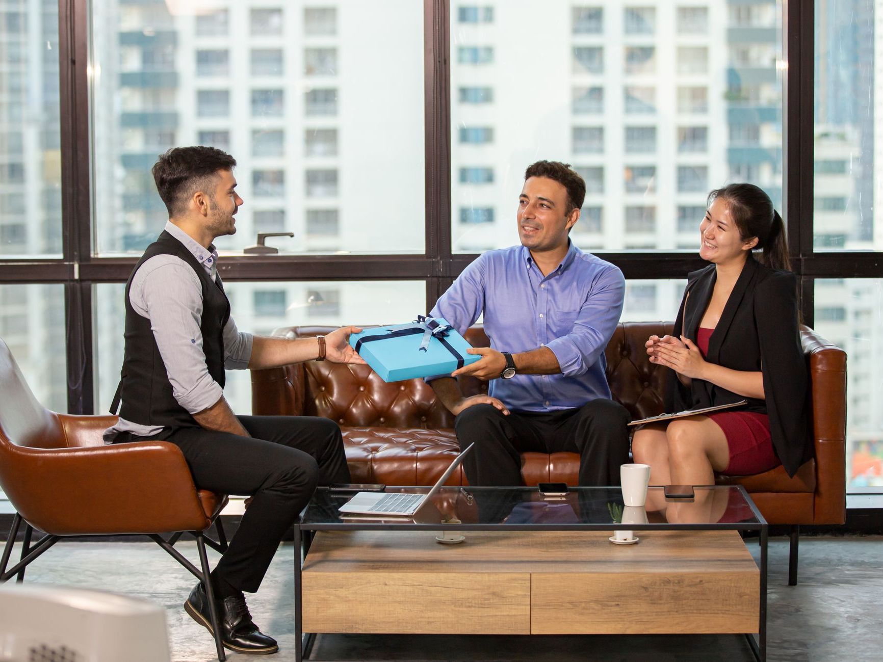 Man in a blue shirt handing a blue gift box to a colleague in a modern office, with a woman beside them smiling.