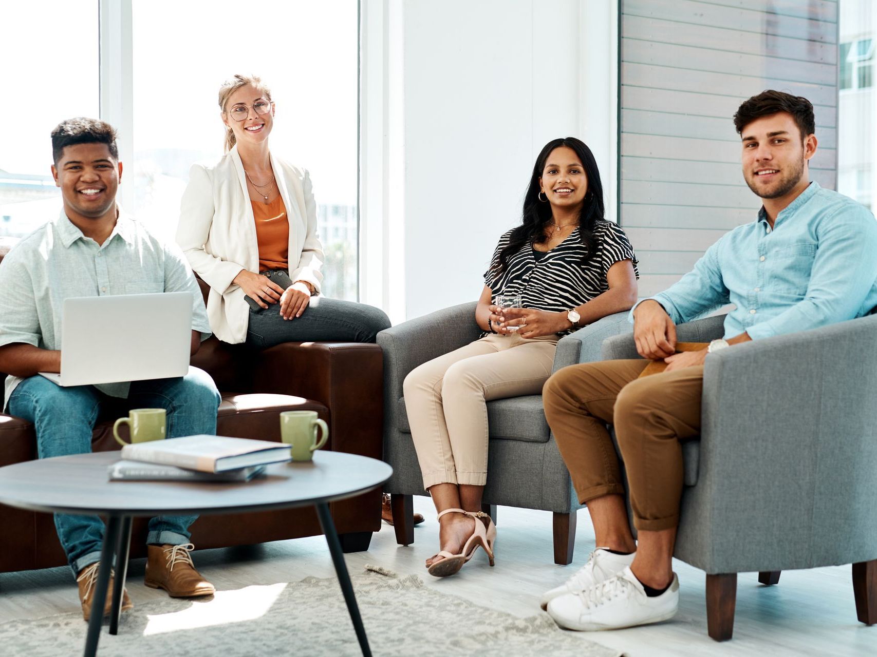 Four professionals sitting in a modern office lounge, smiling and relaxed, with a laptop and coffee mugs on the table.