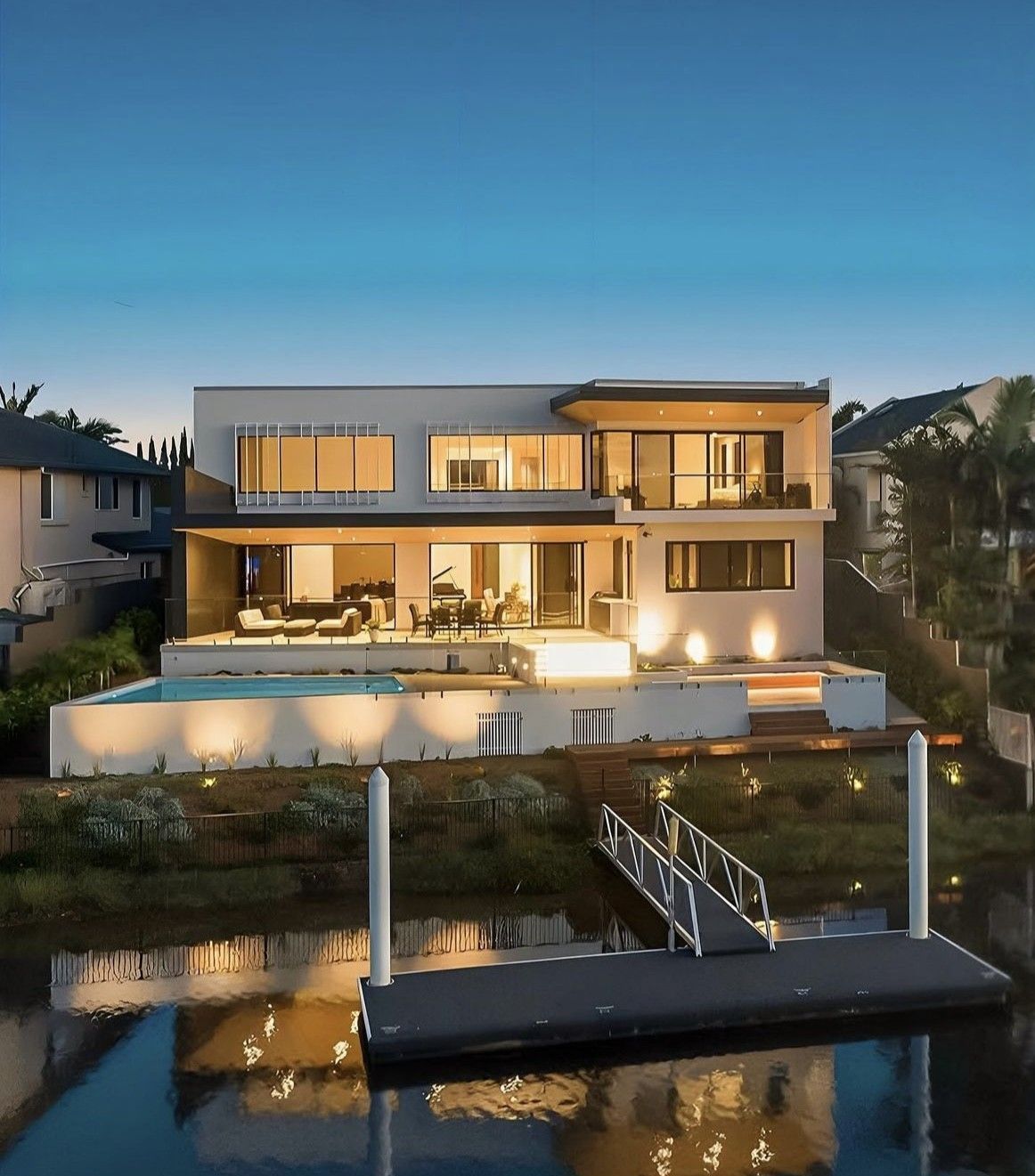 Modern waterfront home with a pool, dock, and evening lights reflecting in the water.