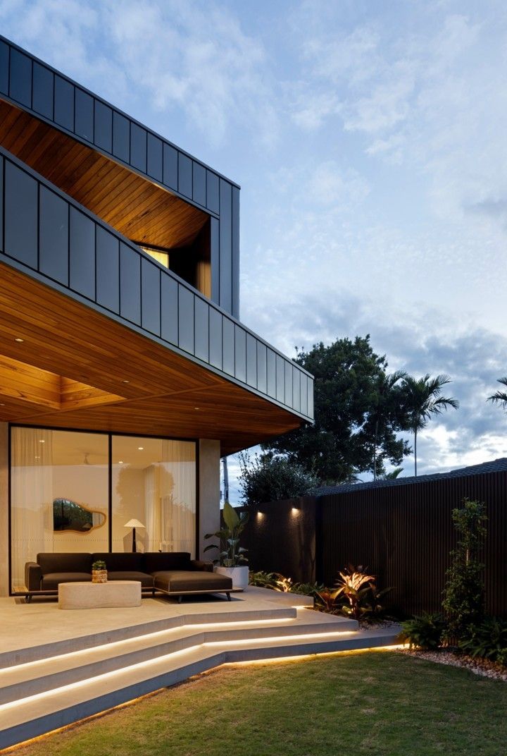 Modern two-story house with warm interior lights, wooden overhang, and a landscaped front lawn at dusk