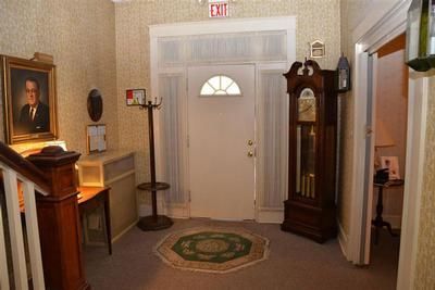 Hallway with white front door, grandfather clock, framed portrait, small table, and green rug