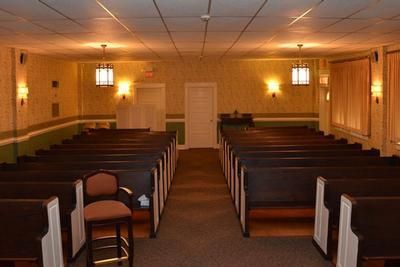 Empty chapel interior with wooden pews, warm wall sconces, and a central aisle leading to a door