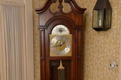 Wooden grandfather clock with a gold clock face in a patterned wallpapered room.