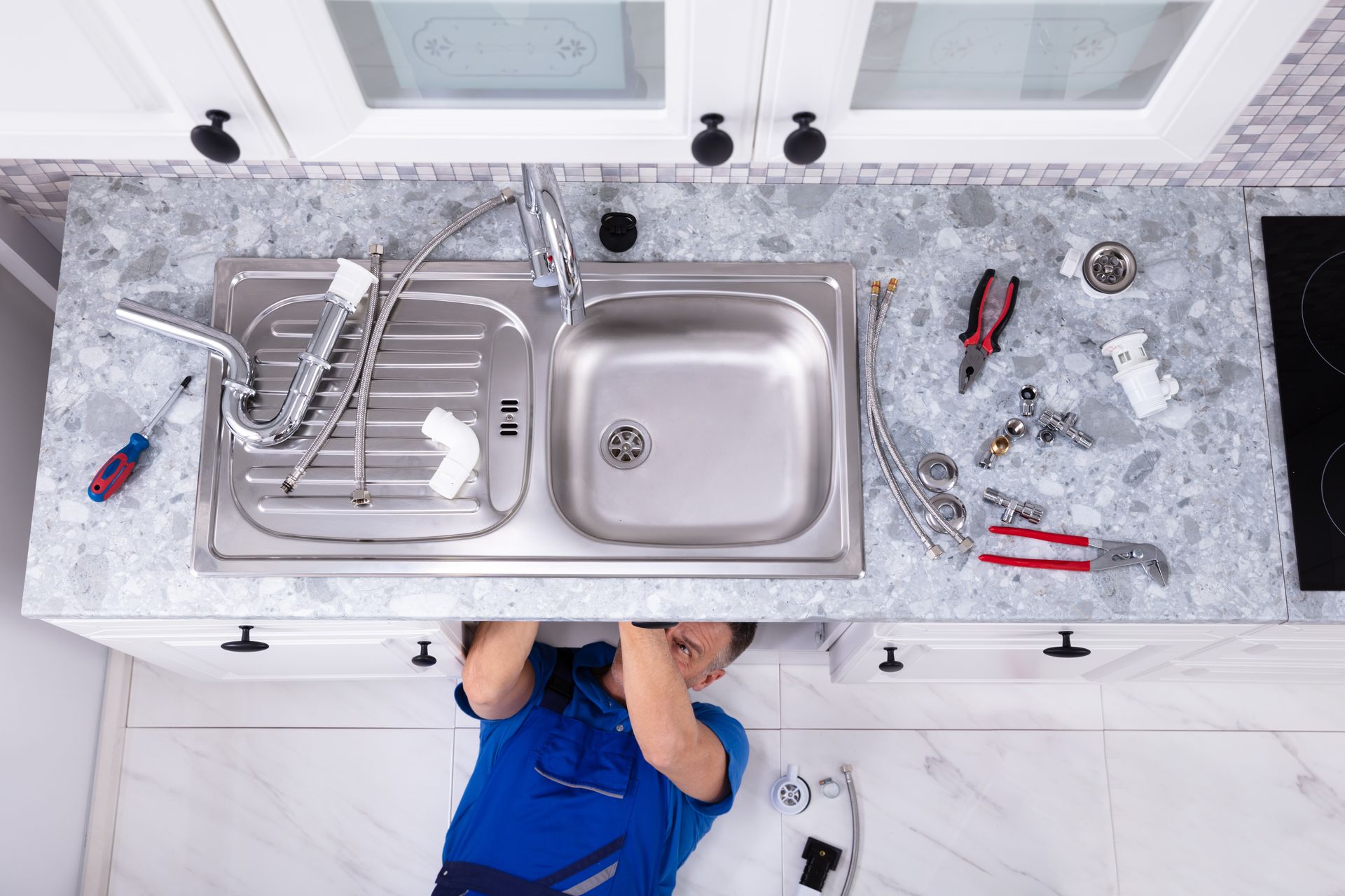 A plumber is working under the kitchen sink with a faucet and tools.
