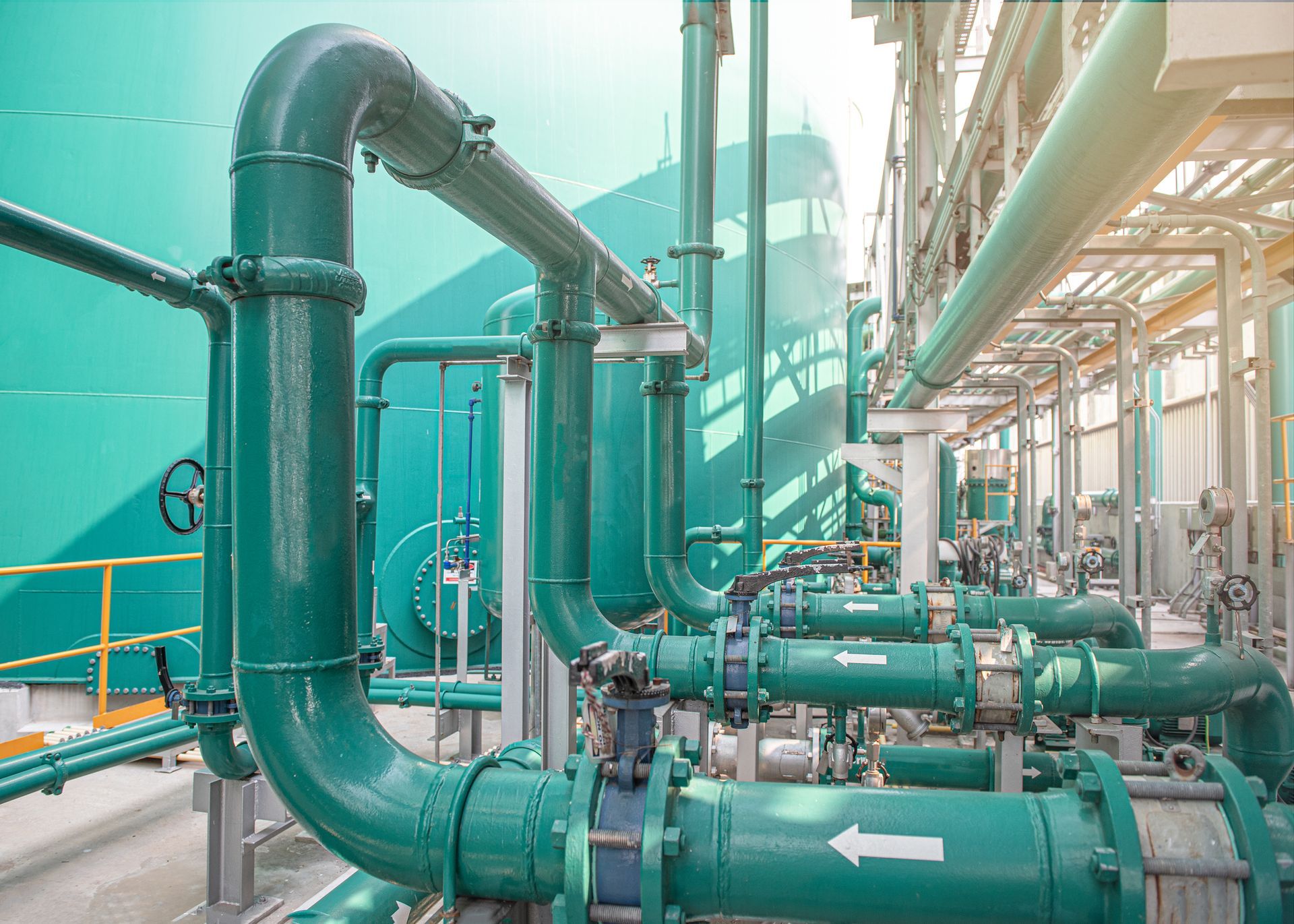 Green industrial pipes and valves in a facility with a large teal tank in the background.