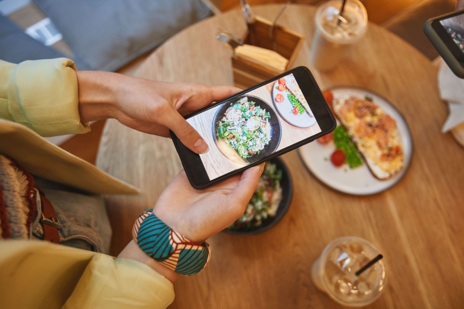 Person using a smartphone to photograph a meal on a wooden restaurant table, with drinks nearby