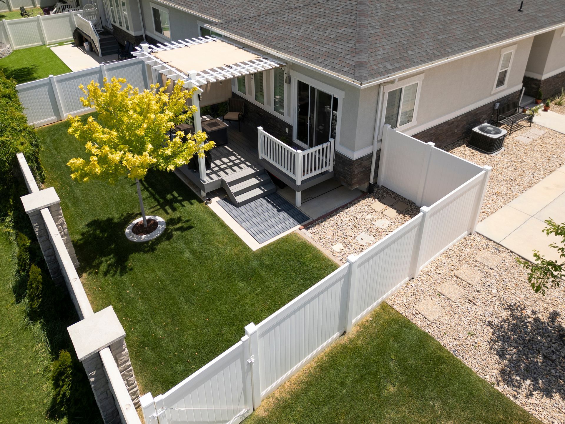 Aerial view of a backyard with green grass, a white fence, patio, and a tree.