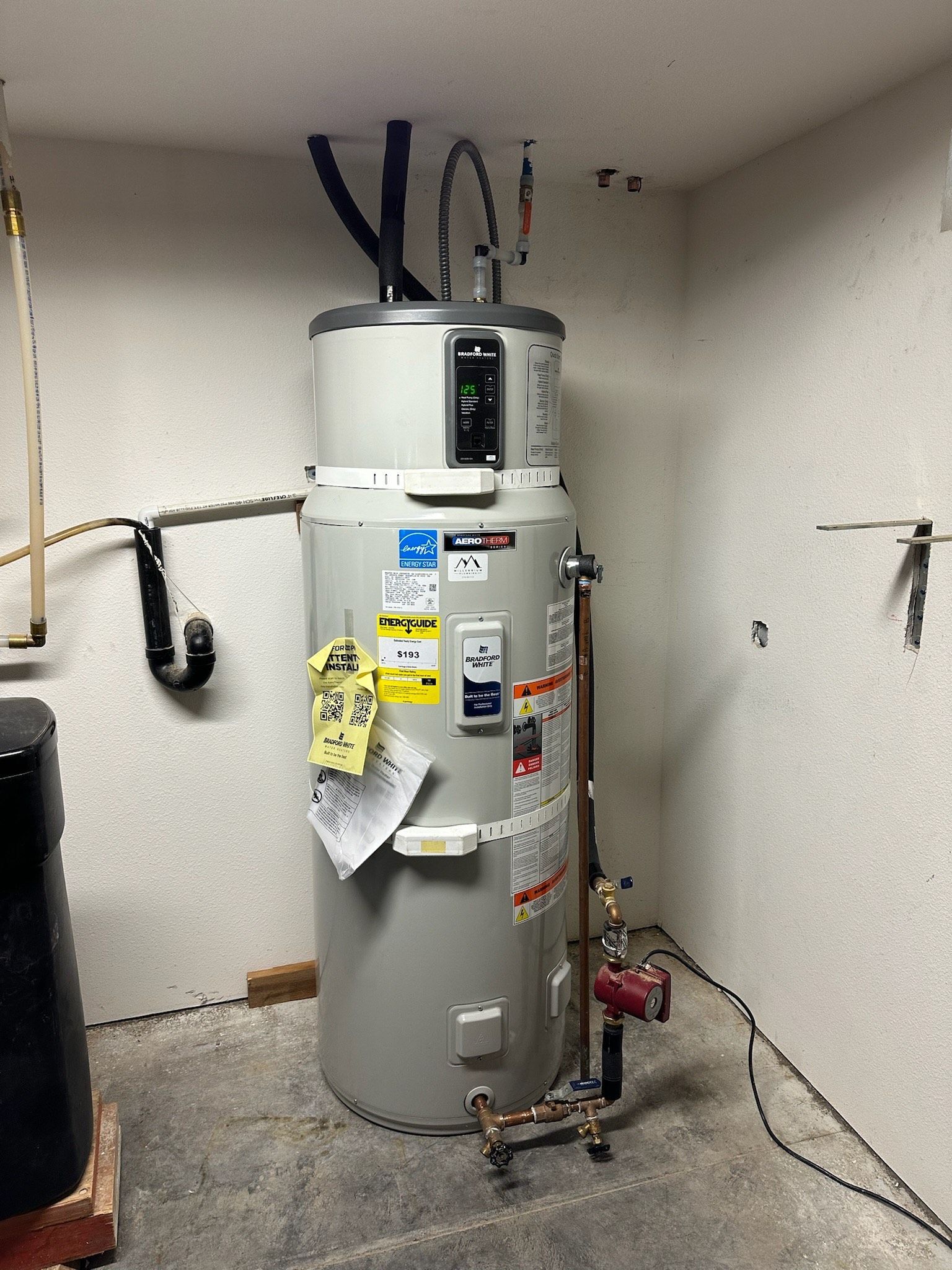 A tall, gray heat pump water heater with connected copper piping, wiring, and labels in a basement corner.