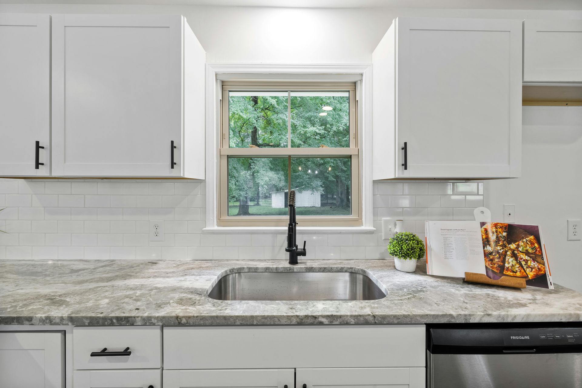 A kitchen sink with a black faucet, granite countertops, white cabinets, and a window overlooking greenery.