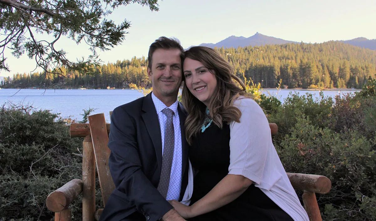 A couple sits together on a wooden bench outdoors, smiling in front of a lake and forest landscape.