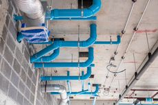 Blue PVC pipes and flexible silver ventilation ducts are suspended from a concrete ceiling in a construction site.