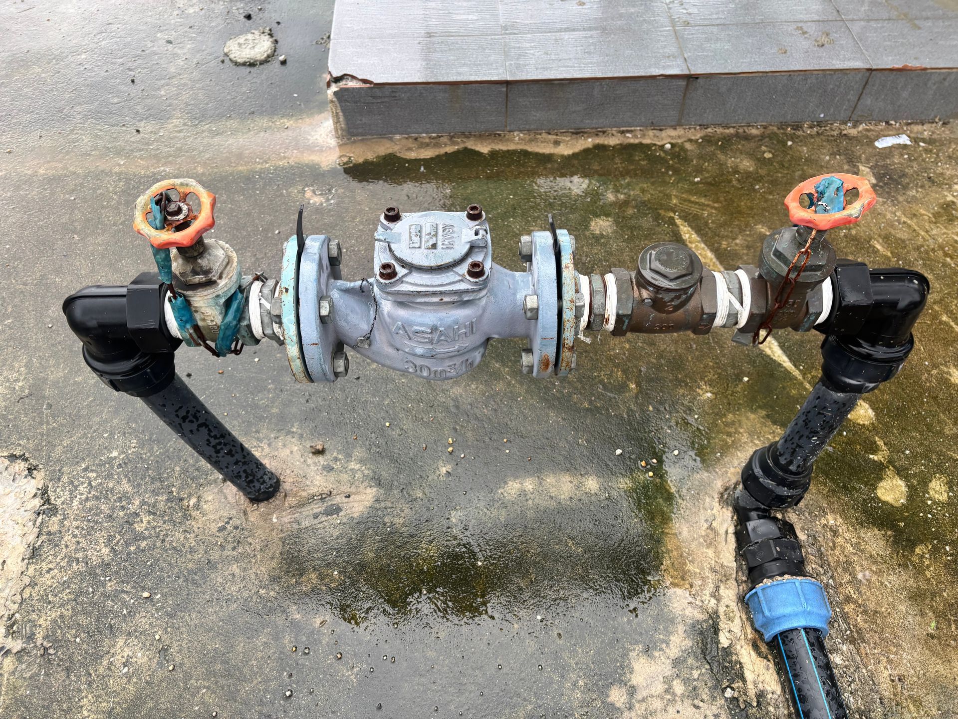 A water meter assembly with two orange-handled valves, pipes, and fittings, situated on a concrete ground surface.