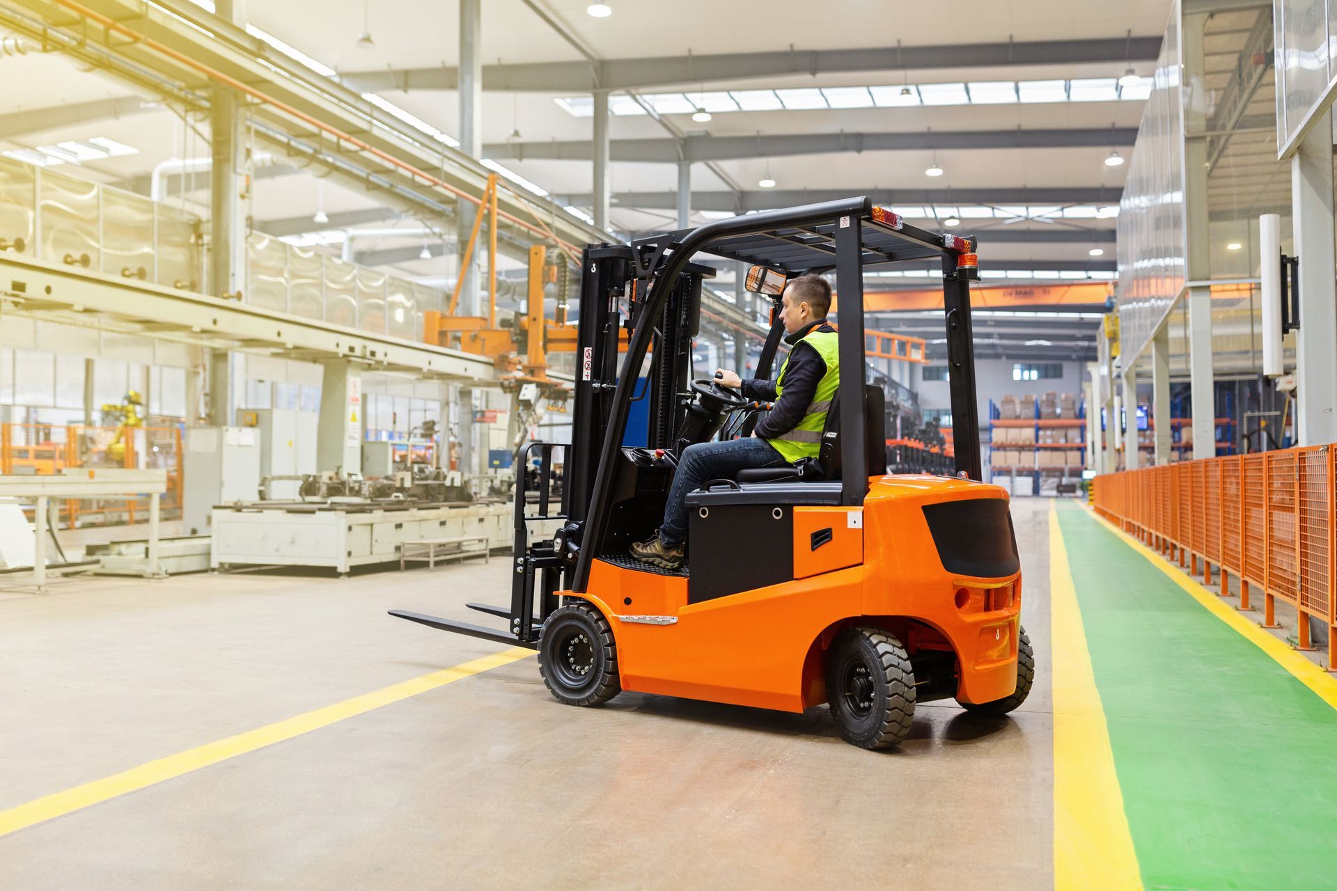 Orange forklift driving through a warehouse aisle beside orange shelving and a green floor lane