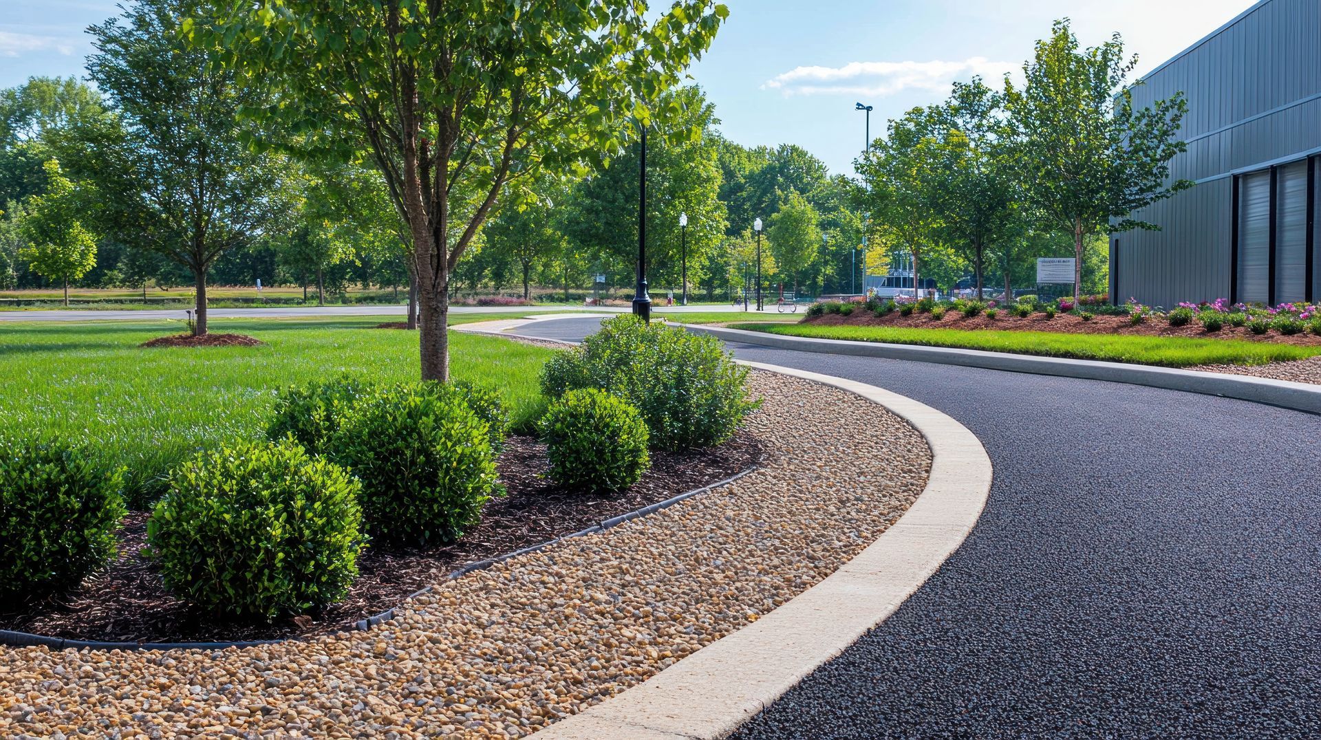 Curved landscaped driveway beside a modern building, with trees, shrubs, and mulch beds in bright daylight