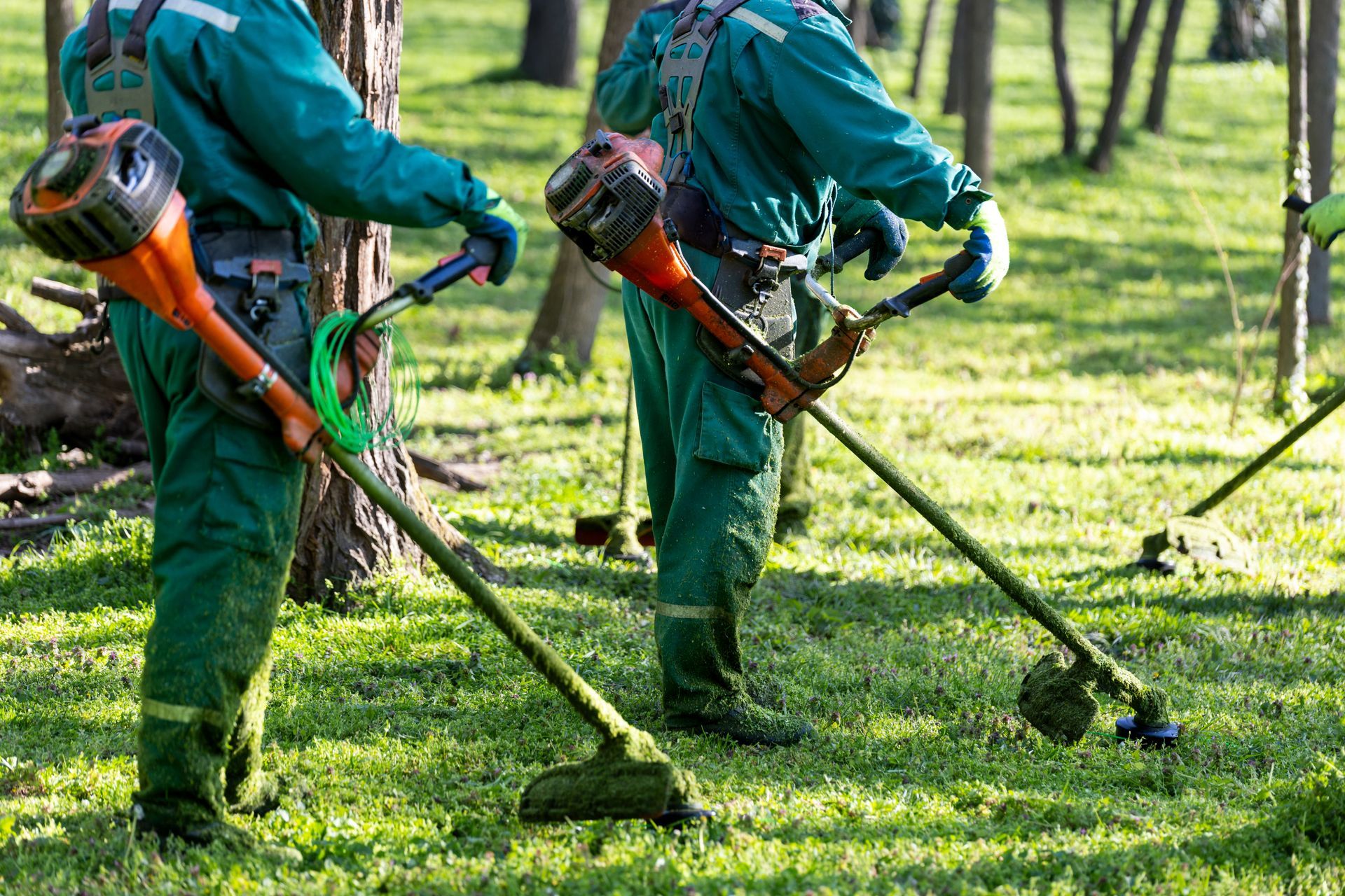 Two landscapers in green uniforms using string trimmers to cut grass in a sunny park