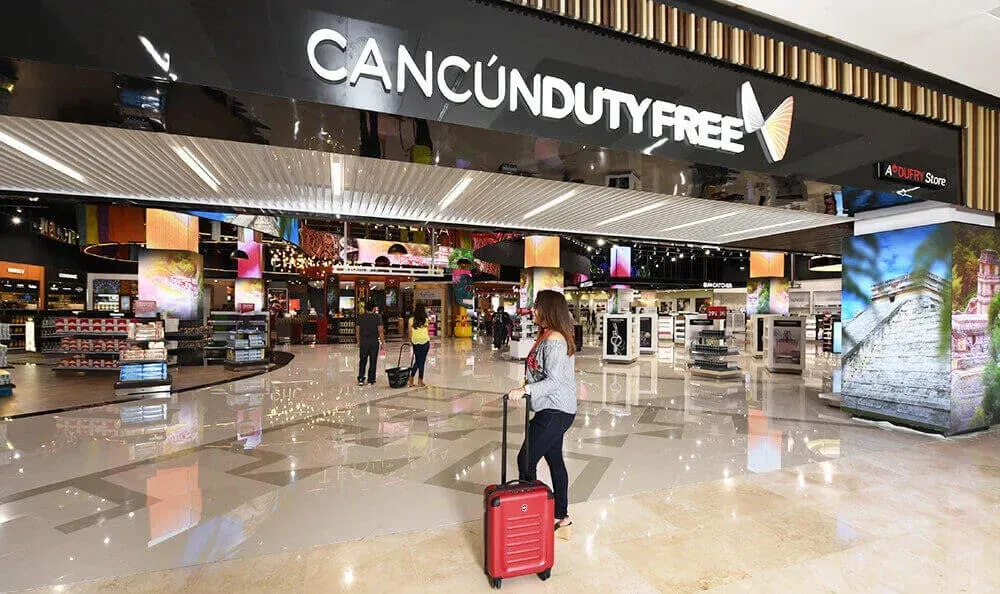 Un viajero con una maleta roja camina hacia la entrada de una tienda libre de impuestos de Cancún, ubicada dentro de una terminal de aeropuerto moderna y luminosa.
