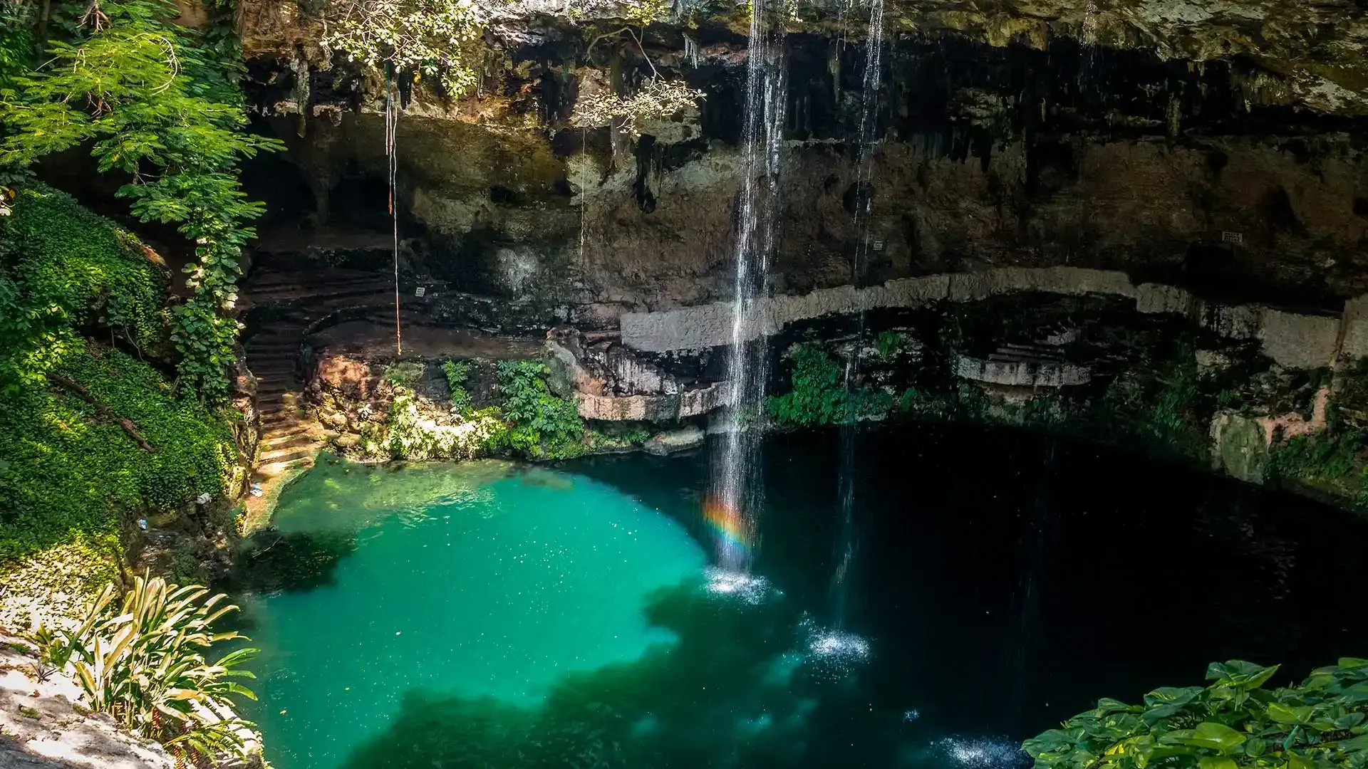 Un sumidero circular y exuberante con una fina cascada que desemboca en una profunda poza de color turquesa rodeada de paredes de piedra caliza.