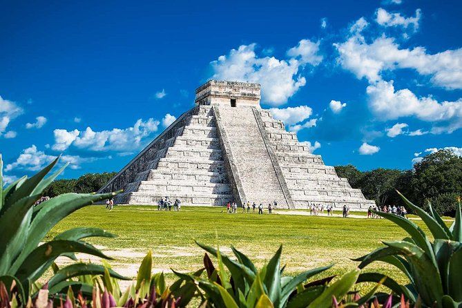 La pirámide de piedra de El Castillo en Chichén Itzá se alza bajo un cielo azul brillante, rodeada de hierba y plantas en primer plano.