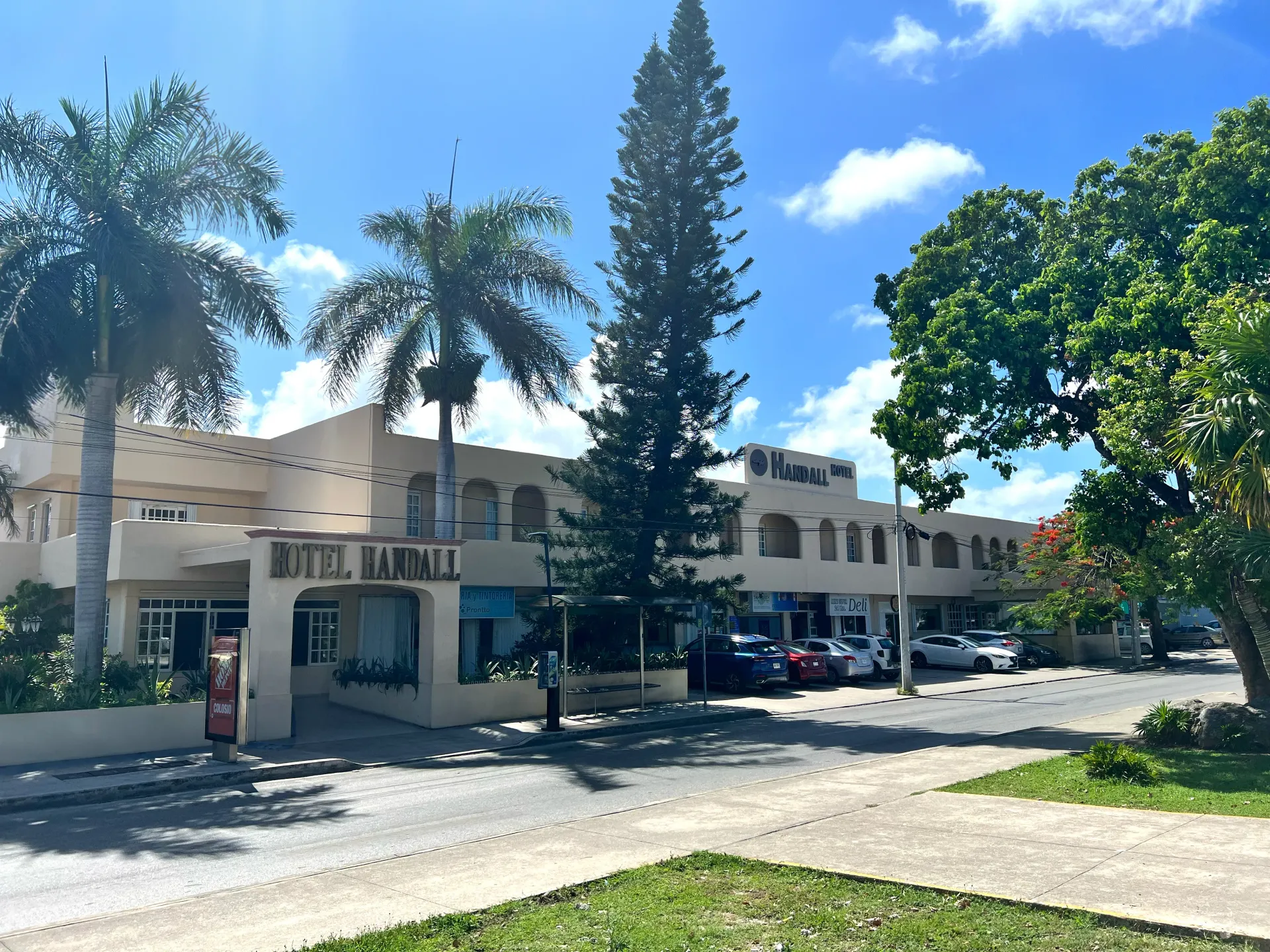 Exterior del Hotel Bahía, un edificio beige de varias plantas con palmeras y una zona de aparcamiento pavimentada bajo un cielo azul brillante.