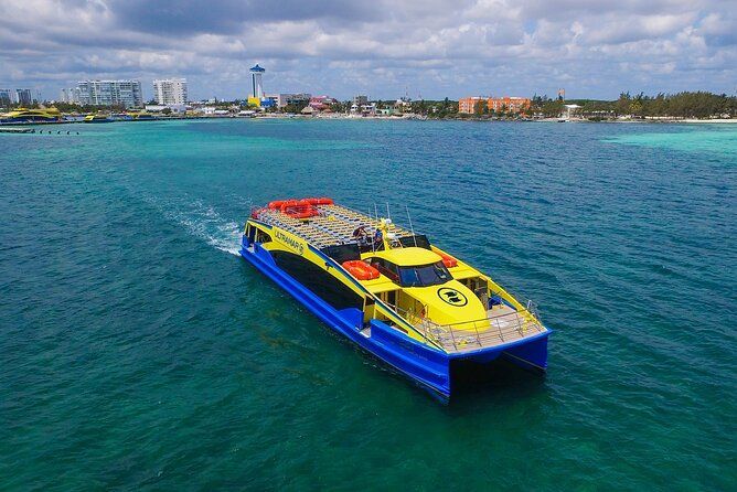 Un catamarán amarillo y azul navegando por aguas tropicales de color turquesa hacia una ciudad costera lejana.