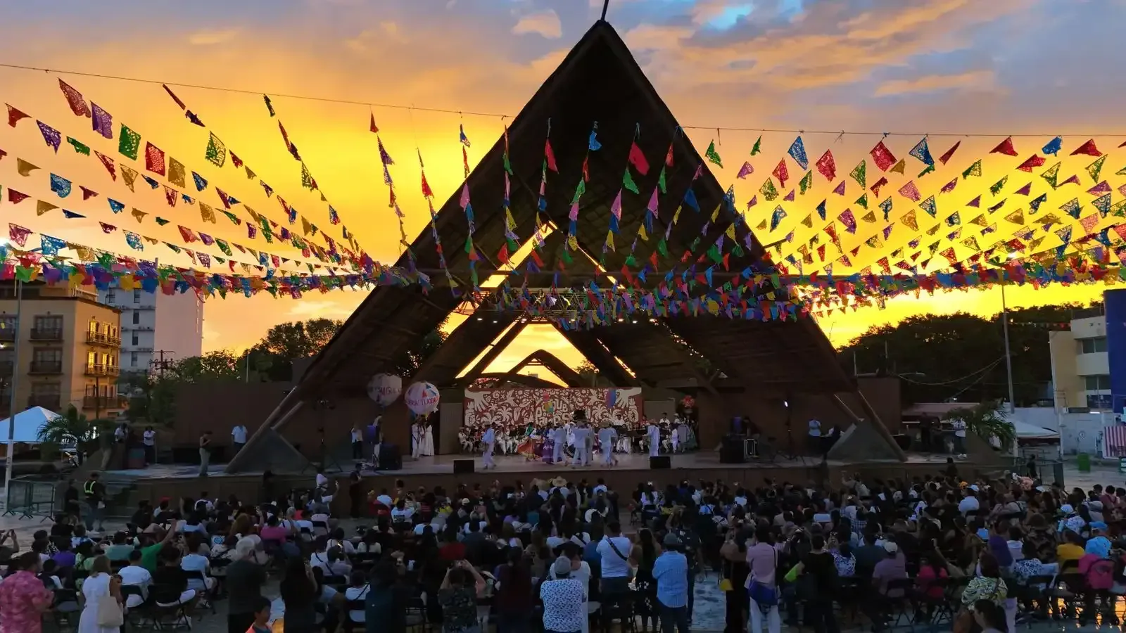 Al atardecer, una multitud observa una actuación en un escenario situado bajo una gran estructura triangular de madera, decorada con coloridas banderas.