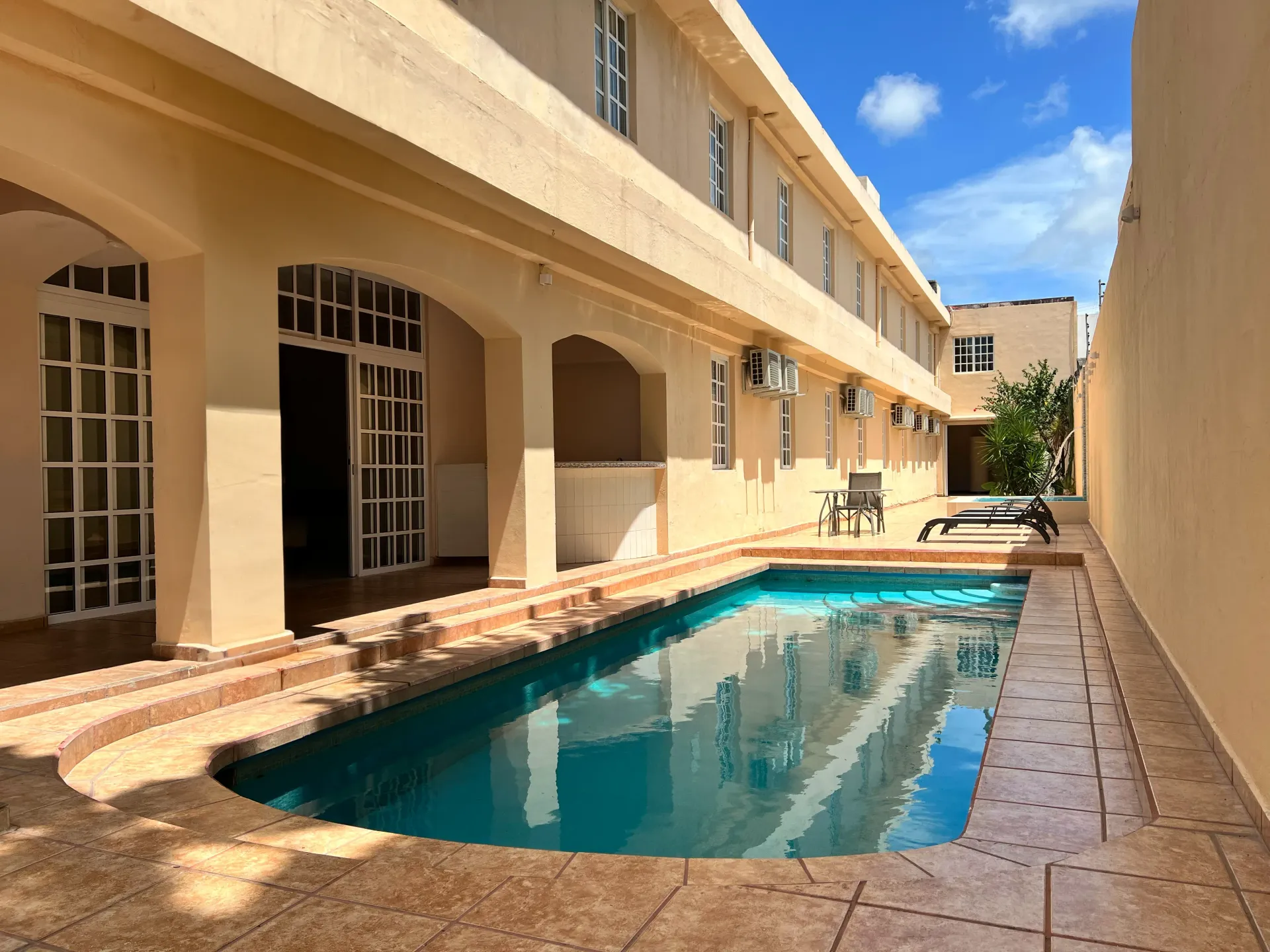 Una piscina rectangular al aire libre, bordeada por un edificio de color beige con puertas arqueadas y una terraza de baldosas a juego.