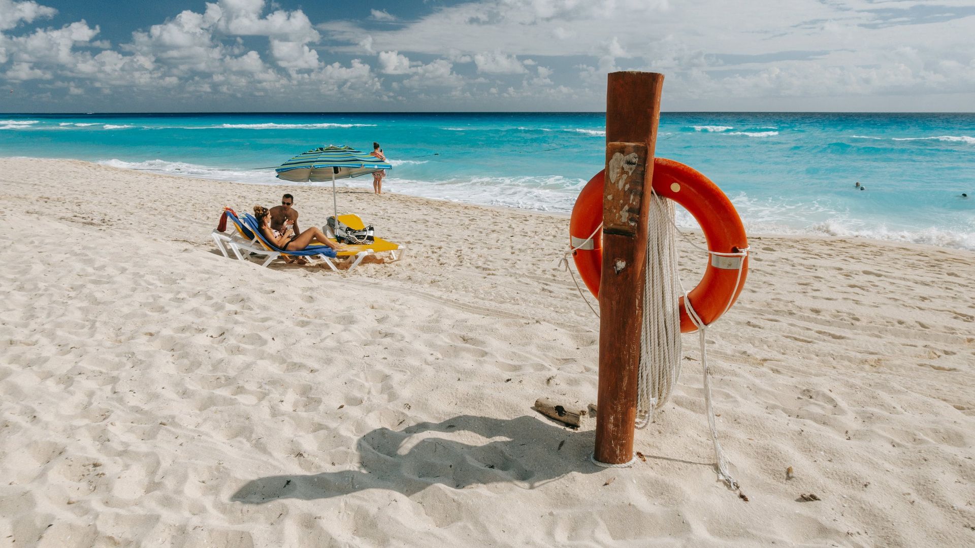 Un salvavidas naranja cuelga de un poste de madera en una playa de arena, con gente relajándose en tumbonas al fondo.