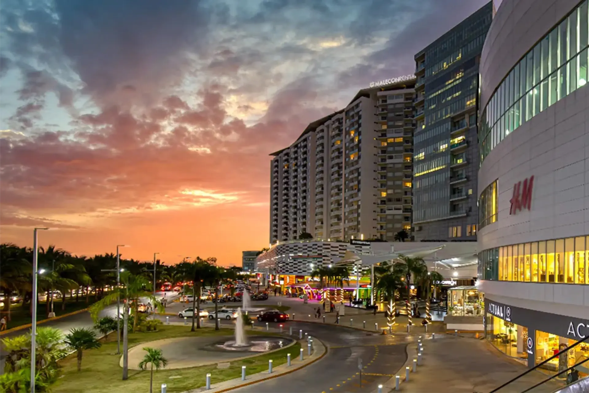 Puesta de sol sobre un moderno centro comercial y un complejo hotelero de gran altura con escaparates iluminados y una entrada ajardinada.