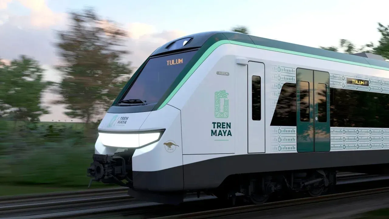 Un tren de pasajeros Tren Maya, de color blanco y verde, avanza a gran velocidad por una vía que atraviesa un paisaje borroso.