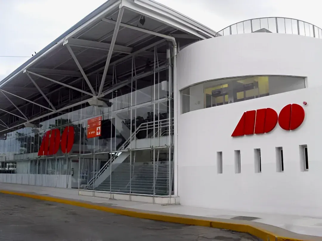 Una moderna estación terminal de autobuses ADO de color blanco, con fachada de cristal y el logotipo rojo de la empresa, en un día nublado.