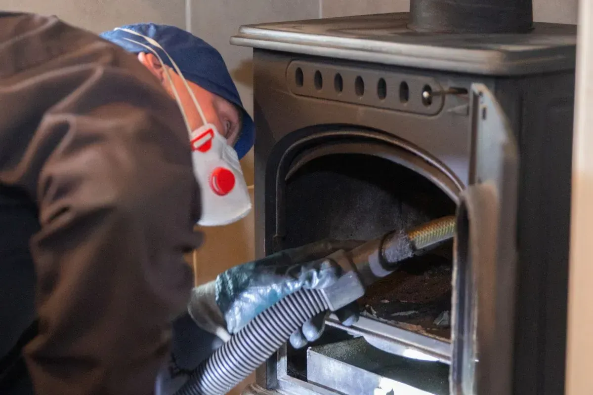 Chimney Sweep Portland Maine-Person in mask and gloves vacuuming a wood stove to remove ash and debris.