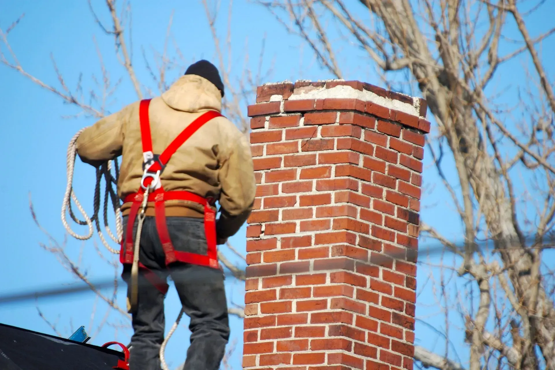 Chimney Sweep Portland Maine-A worker in a brown jacket and red harness stands on a roof, holding a rope next to a red brick chimney. Leafless trees and a clear blue sky in the background.