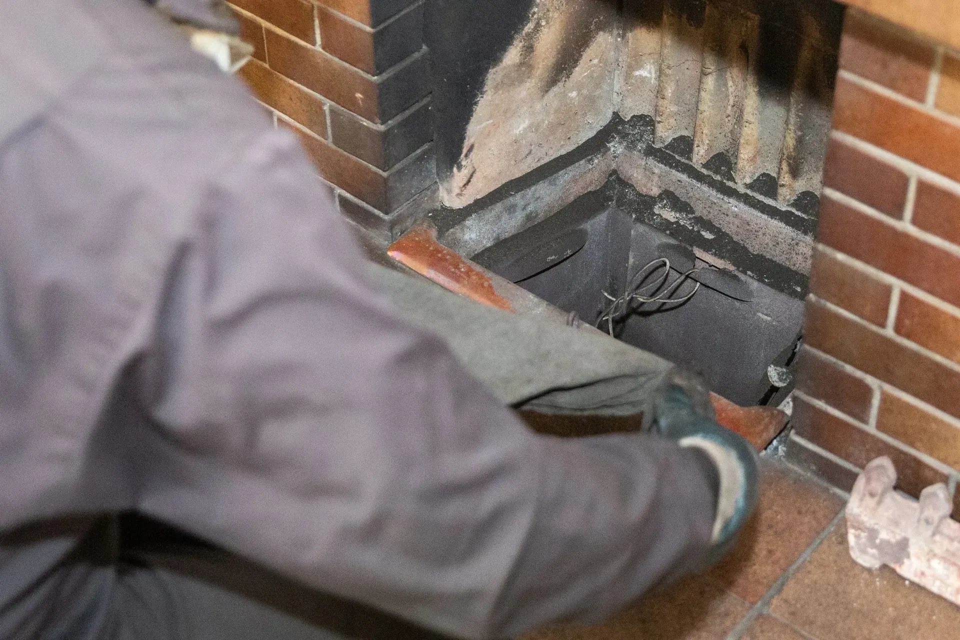 Chimney Sweep Portland Maine-A person cleans a brick fireplace, wearing a grey jacket and gloves, focused on removing ash and debris. The atmosphere conveys maintenance and care.