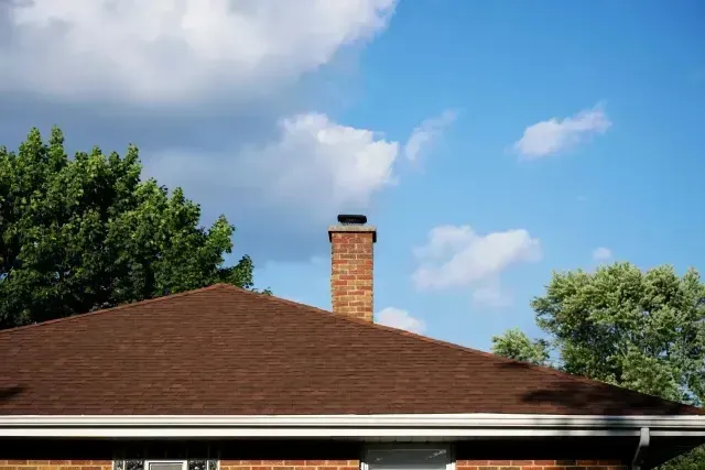 Chimney sweep portland maine - Brown shingled roof with a brick chimney against a bright blue sky with scattered white clouds and trees.