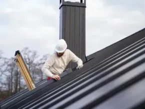 Chimney sweep portland maine - Roofer in hard hat works on a dark metal roof near a chimney.