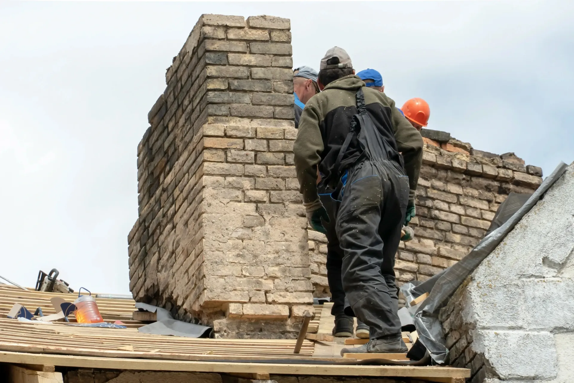 Chimney sweep portland maine - Construction workers in safety gear work on a brick chimney repair on a rooftop with overcast skies.