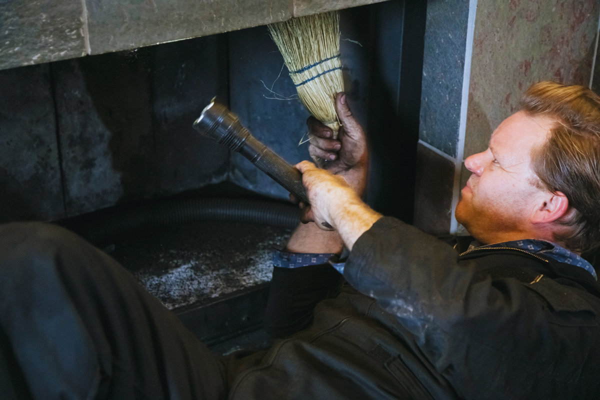 Person cleaning a fireplace with a broom and vacuum, inside a dark stone hearth.