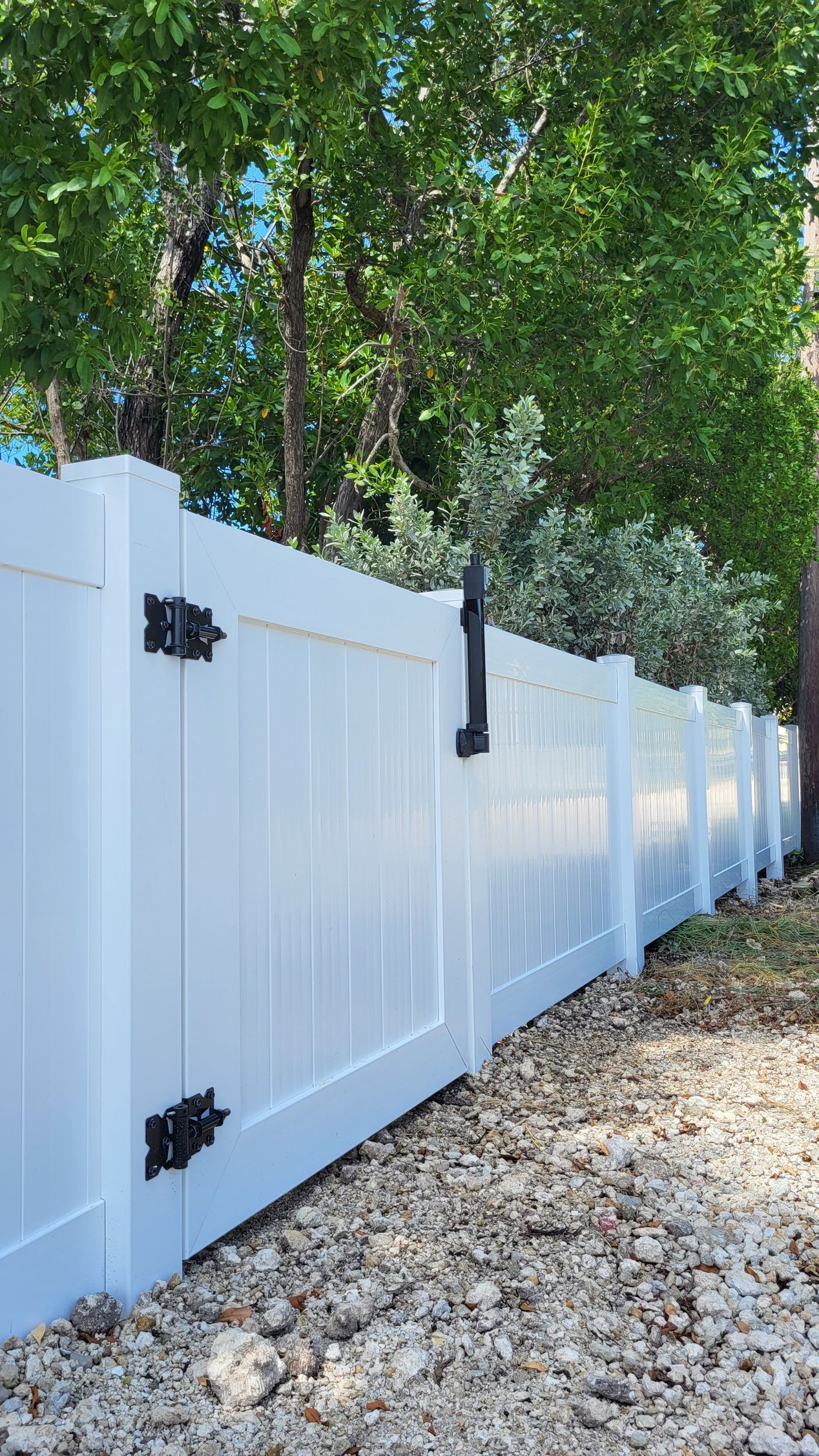 White vinyl fence with gate, black hardware. Green trees in background.