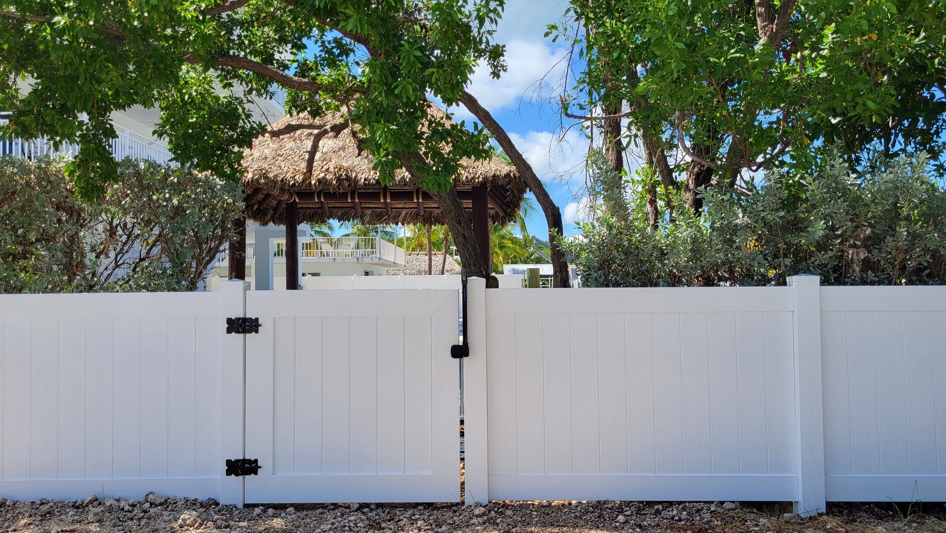 White fence with gate leading to a shaded structure under trees with blue sky visible.