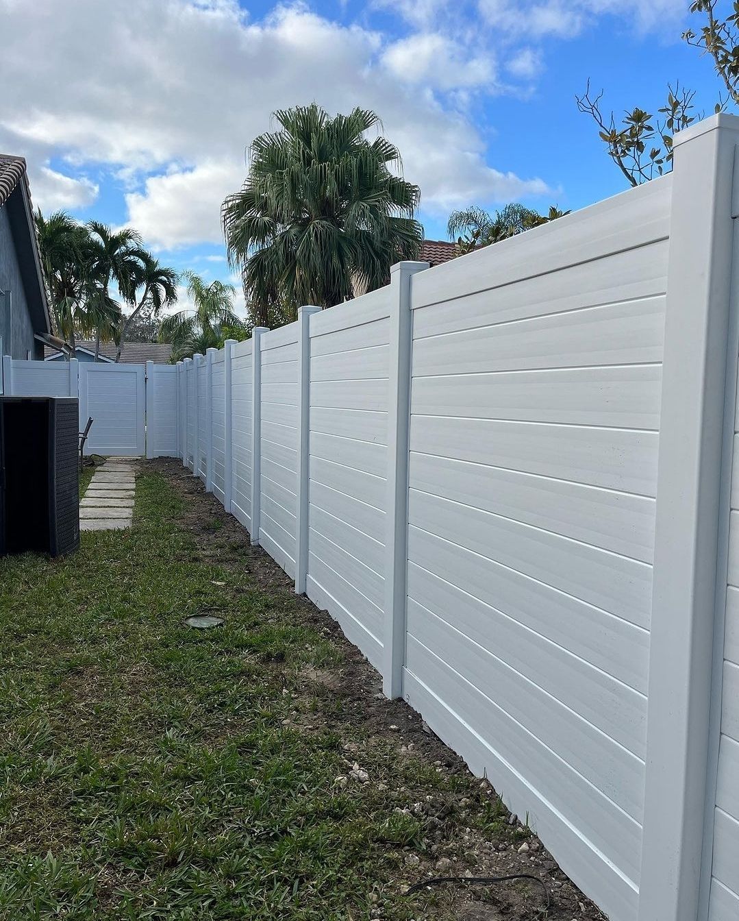 White horizontal-slat privacy fence in backyard with patchy grass. Cloudy sky and palm trees in the background.
