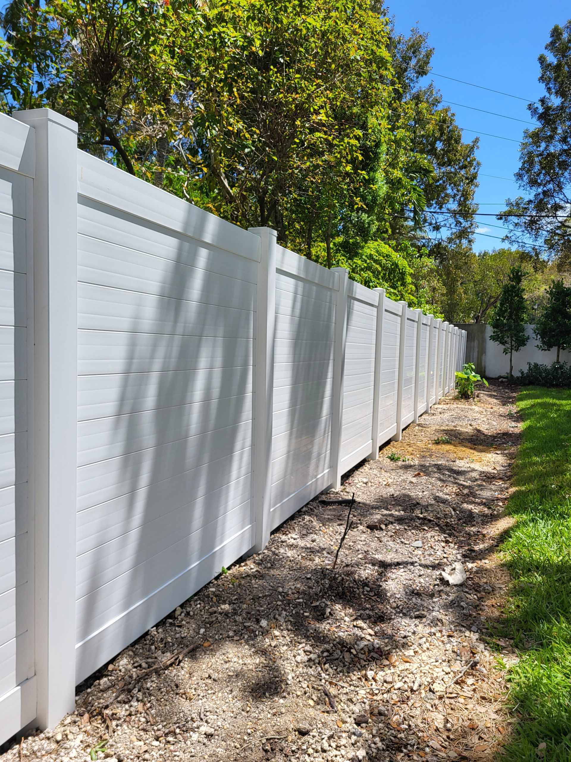 White horizontal slat fence in a yard with gravel and green grass. Blue sky and trees in the background.